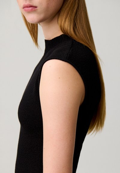 Giovane con lunghi capelli castani indossa una canotta nera testurizzata senza maniche, rivolto di lato su uno sfondo chiaro e uniforme.