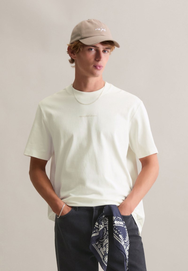 Biała bawełniana koszulka z okrągłym dekoltem, w połączeniu z granatowymi spodniami i niebieską bandaną zawiązaną w talii. Do tego beżowa czapka i srebrny łańcuszek.