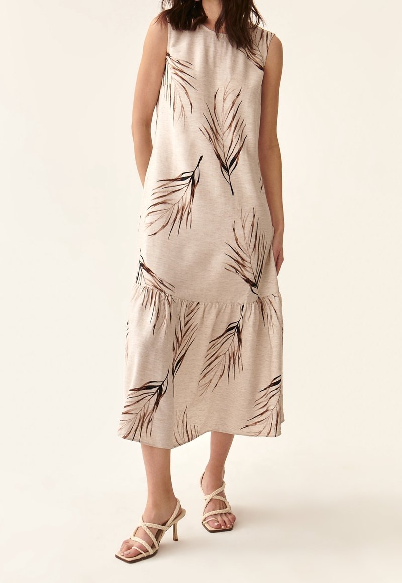 Robe midi beige sans manches à imprimés de feuilles brunes, jupe à volants, portée avec des sandales à talons hauts à brides tressées couleur crème sur fond clair.