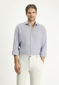 Camicia azzurro chiaro con un motivo testurizzato, maniche lunghe arrotolate fino ai gomiti e colletto classico, abbinata a pantaloni beige.