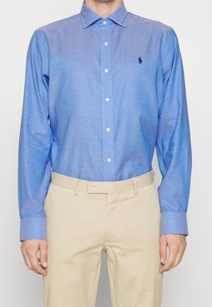 Mężczyzna w jasno-niebieskiej koszuli zapinanej na guziki z drobnym wzorem w kropki i beżowych spodniach, stoi na jednolitym tle.