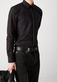 Camisa negra de manga larga con cierre de botones en el frente, que presenta un zipper en contraste a lo largo del pliegue. Combinada con jeans negros y un cinturón negro.