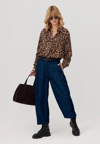 Kobieta w bluzce w panterkę, szerokich niebieskich spodniach, czarnych botkach, w okularach przeciwsłonecznych i trzymająca dużą ciemnobrązową torebkę na jednolitym tle.