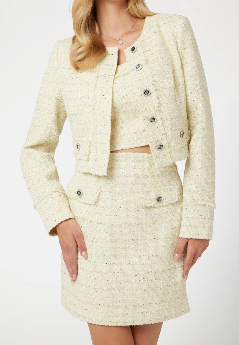 Femme portant une tenue deux pièces texturée crème avec une veste courte, un top sans manches et une jupe taille haute, tous avec des détails de boutons.