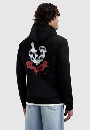 Man met krullend haar die een zwarte hoodie draagt met een witte en rode gestippelde handafbeelding en de tekst "pura path" op de achterkant, gecombineerd met lichtblauwe spijkerbroek.