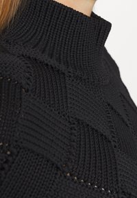 Sort strikket sweater tæt på med en høj krave og strukturelle mønstre. Stof har et ribbet design og en glat overflade.