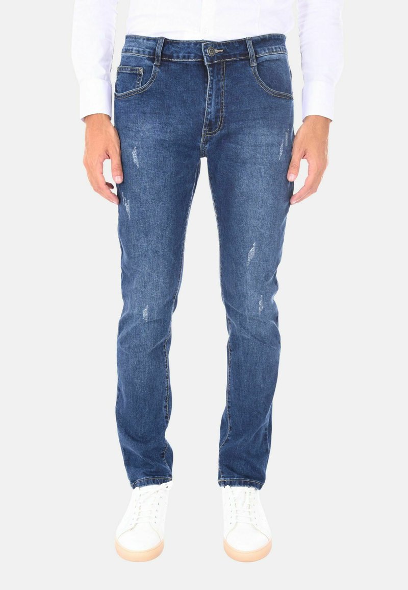 Jeans in denim blu dal taglio slim-fit con leggere rotture e cinque tasche, indossati con sneaker bianche e una camicia a maniche lunghe.
