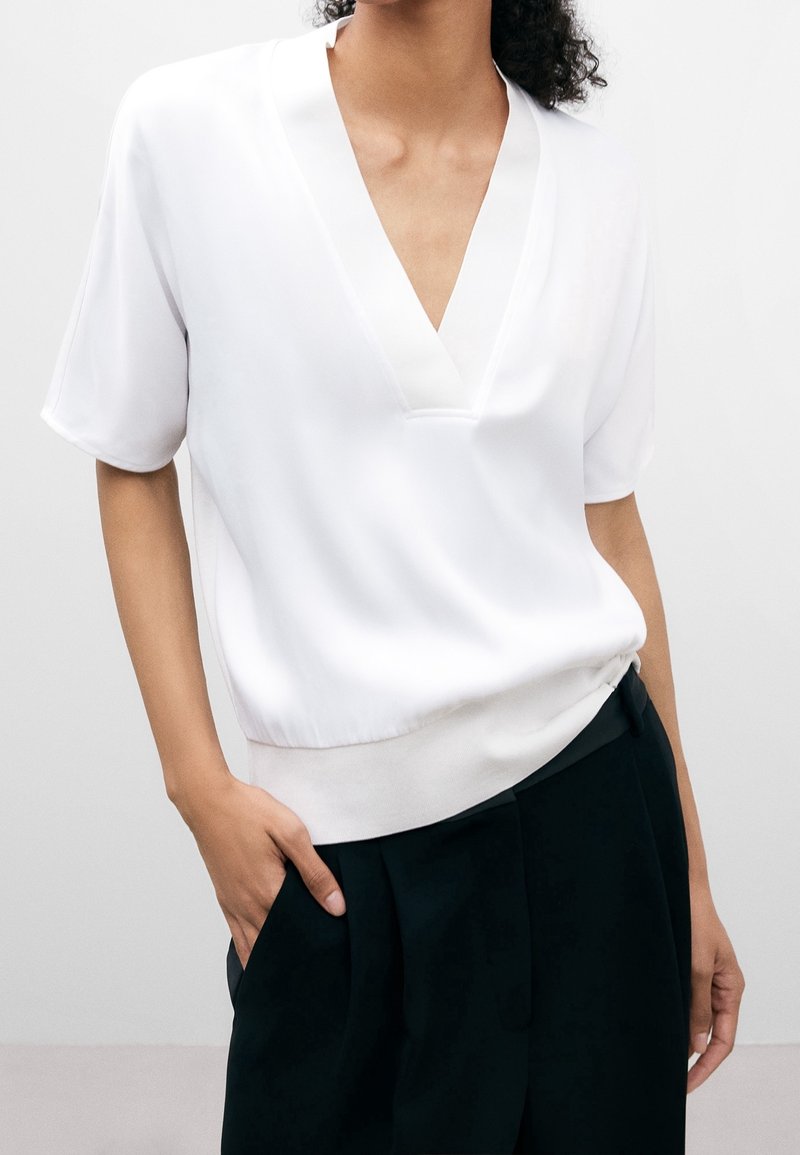 Blouse blanche à col en V en tissu lisse, dotée de manches courtes et d'une coupe décontractée, agrémentée d'une ceinture douce et contrastante.