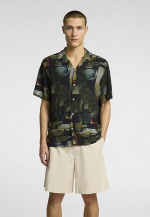Camicia a maniche corte, con bottoni, realizzata in un tessuto scuro a fantasia, con forme astratte nei toni del verde, marrone e arancione, abbinata a pantaloni corti beige chiaro.
