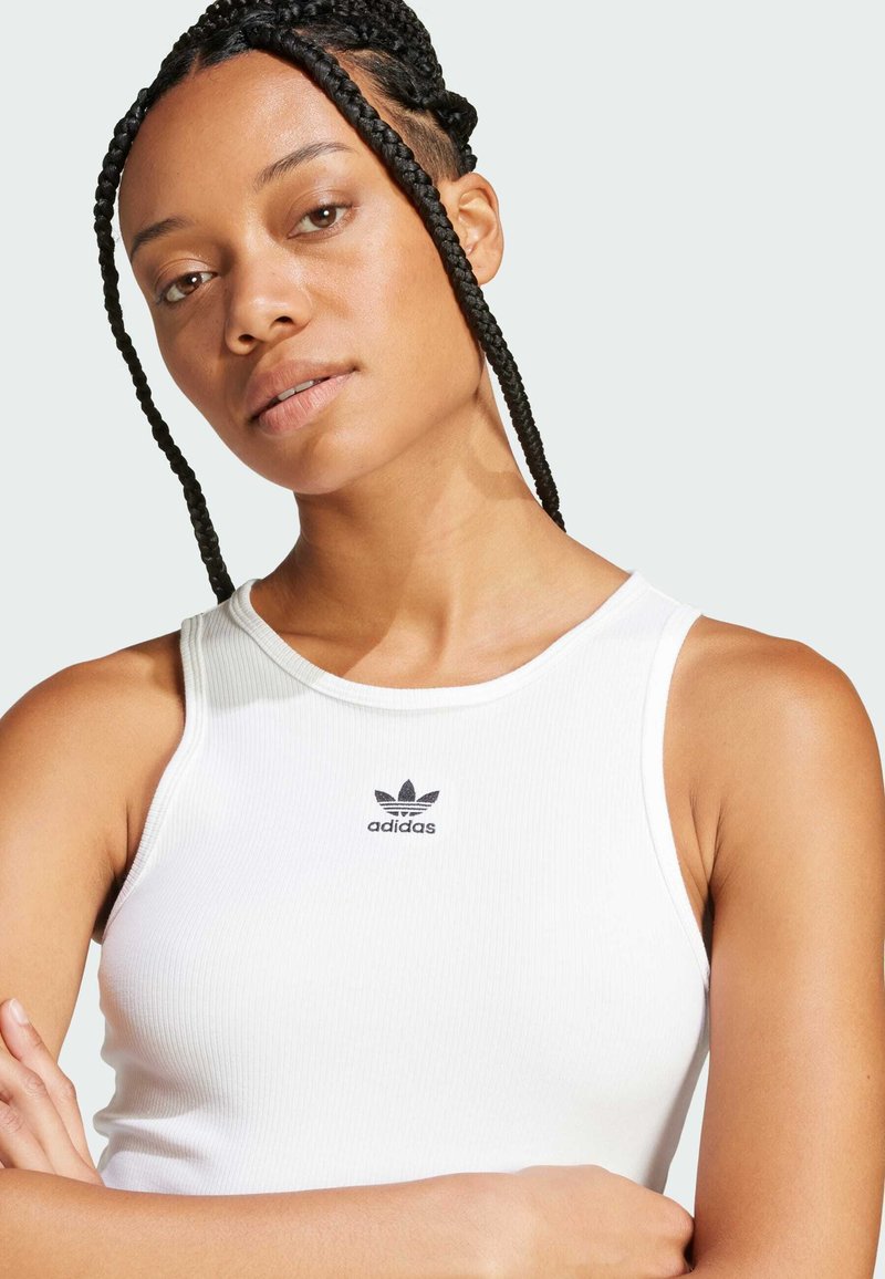 Vit ribbad tanktop med rund halsringning och en liten svart Adidas-logotyp på bröstet. Slät textur och figurnära design.