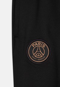 Los pantalones de chándal negros presentan un parche bordado circular en bronce con el texto "PARIS" y "SAINT-GERMAIN", acentuando el diseño.