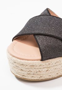Czarne brokatowe klapki na platformie z plecionki jutowej, z krzyżującymi się paskami i gładką beżową wkładką z widocznym przeszyciem.