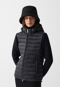 Zwarte puffer vest met horizontaal doorgestikte patroon, ritssluiting, zijzakken en een capuchon, gecombineerd met een zwarte gebreide trui en een bucket hat.