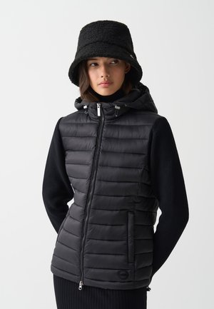 Gilet invernale nero con motivo a righe orizzontali, chiusura con zip, tasche laterali e cappuccio, abbinato a un maglione in maglia nero e un cappello a secchiello.