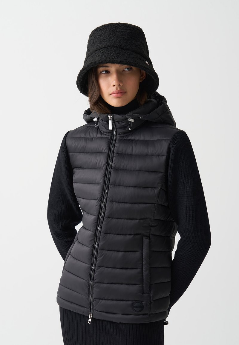 Zwarte puffer vest met horizontaal doorgestikte patroon, ritssluiting, zijzakken en een capuchon, gecombineerd met een zwarte gebreide trui en een bucket hat.