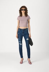 Camiseta morada corta, jeans azules rasgados, bolso negro y tacones morados con tiras. Corte recto con desgastes visibles en los jeans.