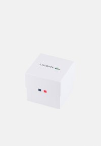 Caja de cartón blanca con tapa, con el logo "LACOSTE", un emblema de cocodrilo verde y pequeños acentos cuadrados rojos y azules en la parte frontal.
