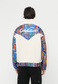 Fleece jacket with a cream body and a colorful patterned fabric on shoulders and sleeves. Features a high collar and logo text on the back.