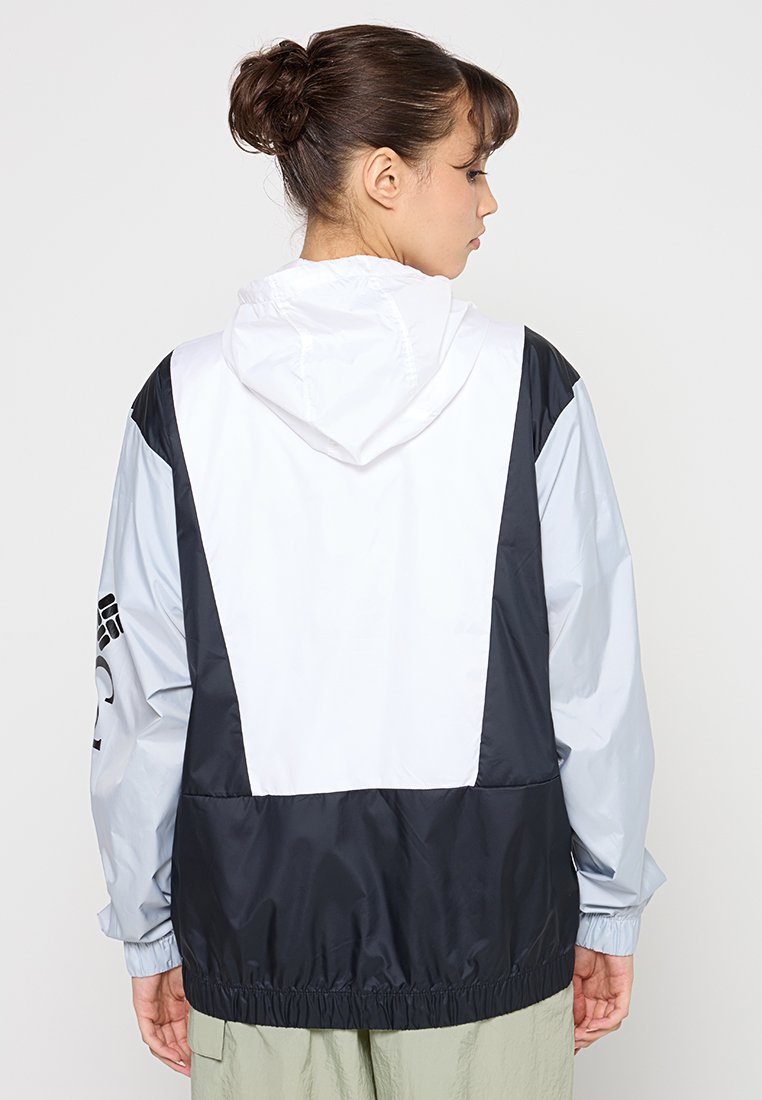 Cazadora tipo windbreaker en blanco, gris claro y gris oscuro. Presenta una capucha, puños elásticos y un dobladillo ajustado. Detalle del logo en la manga.