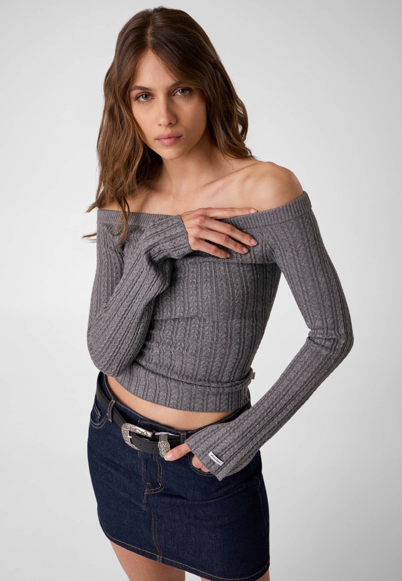Jeune femme portant un pull gris côtelé à épaules dénudées et une mini-jupe en jean foncé avec une ceinture noire, sur fond gris uni.