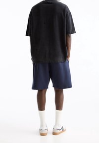 T-shirt noire oversize avec des détails de couture visibles, associée à un short bleu marine. Des chaussettes blanches et des chaussures décontractées complètent le look.