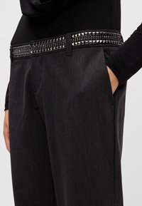 Μαύρο παντελόνι με υφασμάτινη υφή, με ζώνη διακοσμημένη με μεταλλικά καρφιά στη μέση και μια λεία μέση.