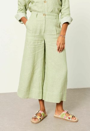 Woman wearing light green wide-leg cropped pants, matching button-up shirt with rolled sleeves, and beige embroidered double-strap sandals.