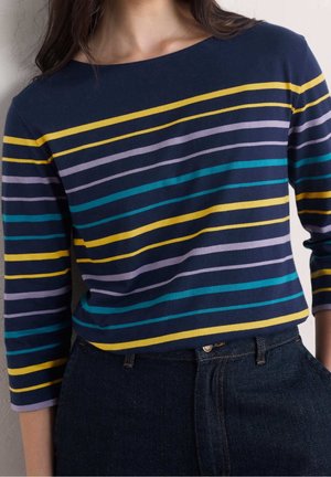 Personne portant une chemise à manches longues bleu marine avec des rayures horizontales jaunes, bleu sarcelle et violet clair rentrée dans un jean bleu foncé.