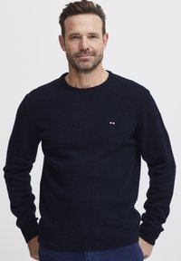Tengerészkék kötött pulóver kerek nyakkal, bordás mandzsettákkal és szegéllyel, a mellkason egy kis piros, fehér és kék logóval. Puha tapintású.