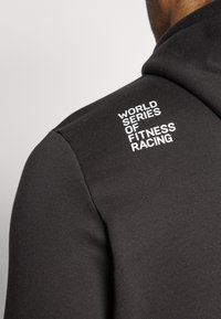 Haut à capuche noir avec une texture lisse, affichant un texte blanc qui dit "WORLD SERIES OF FITNESS RACING" sur l'épaule.