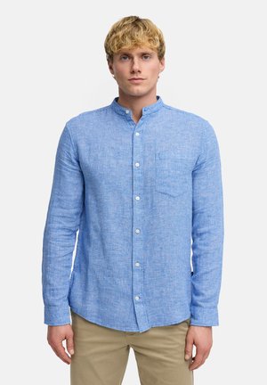 Nuori mies, jolla on vaaleat hiukset ja joka pukeutuu vaaleansiniseen, tekstuuriin mandariinikauluksella olevaan paitaan ja beigen värisiin housuihin, seisoo yksivärisen taustan edessä.