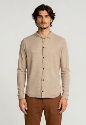 Jonge man met donker haar, draagend een lichtbeige blouse met lange mouwen en knopen en een bruine broek, kijkend naar voren tegen een effen achtergrond.