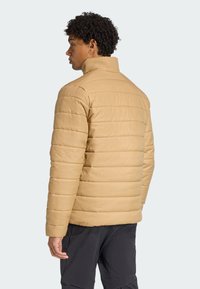 En person med krøllet hår, iført en tan-farvet quiltet jakke, står med ryggen til mod en lys baggrund. Fokus er på jakken design.