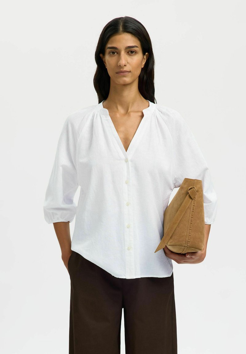 Femme aux longs cheveux bruns portant une blouse blanche boutonnée et un pantalon foncé, tenant un sac à main en daim beige avec une bandoulière dans sa main droite.