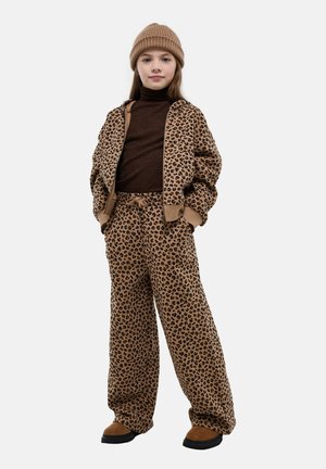 Enfant debout, portant une veste et un pantalon imprimé léopard marron, un pull à col roulé marron foncé, un bonnet beige et des chaussures marron, mains dans les poches.