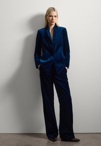 Traje de terciopelo azul marino con un blazer ajustado y pantalones de pierna ancho. La tela tiene una textura suave y el blazer cuenta con un diseño de solapa muesca.