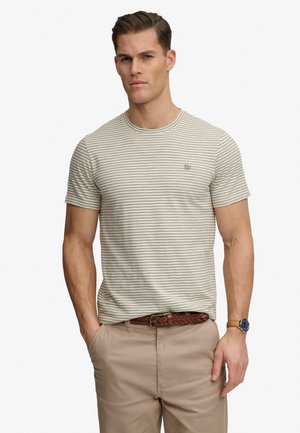 Mann trägt beige gestreiftes T-Shirt, hellbraune Hose mit geflochtenem Gürtel und Armbanduhr, steht mit einer Hand in der Tasche.