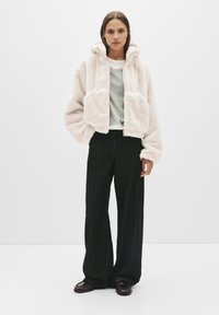 Duks od umjetnog krzna blijedo ružičaste boje, s prednjim patentnim zatvaračem i velikim džepovima. Uparen je s širokim hlačama crne boje s okomitim prugama i tamnom obućom.