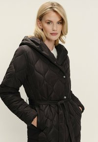 Čierny prešívaný puffa kabát s kapucňou, vybavený opaskom, cvokmi a bočnými vreckami. Elegantná, hladká textúra s diamantom vzorom.