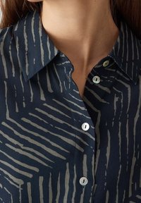 Blusa blu navy con un sottile motivo a righe beige, design con colletto, cinque bottoni e un tessuto morbido e textured. Vista ravvicinata del colletto.