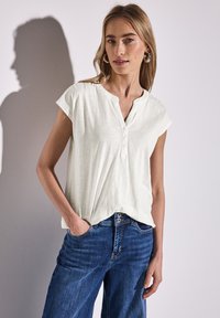 Maglietta corta beige chiaro con scollo a henley e dettagli di bottoni, abbinata a jeans in denim blu. Tessuto morbido con una vestibilità rilassata.