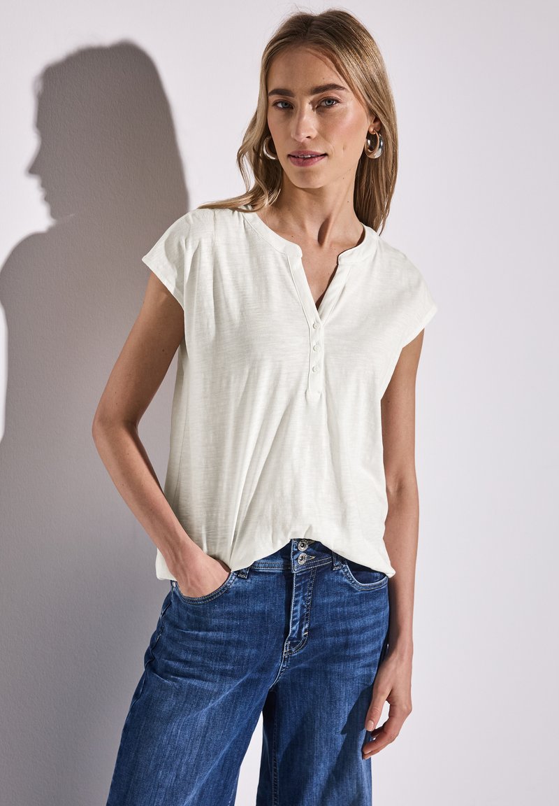 Maglietta corta beige chiaro con scollo a henley e dettagli di bottoni, abbinata a jeans in denim blu. Tessuto morbido con una vestibilità rilassata.