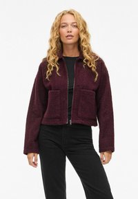 Veste zippée bordeaux en tissu polaire texturé, dotée de deux poches avant et d'un col, accompagnée d'un pantalon noir.
