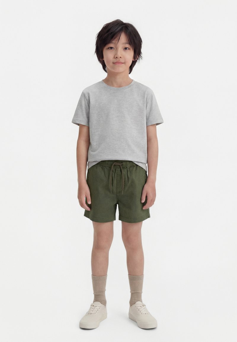 Νεαρός αγόρι στέκεται μπροστά, φοράει ανοιχτό γκρι μπλουζάκι, χακί πράσινα σορτς, μπεζ κάλτσες και λευκά αθλητικά παπούτσια μπροστά σε απλό φόντο.