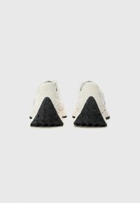 White suede and mesh sneakers with black rubber triangular sole, featuring textured accents on heel and logo detail on side.