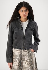 Chaqueta de mezclilla negra recortada con cierre de cremallera, bolsillos frontales y costuras en contraste. Llevada sobre pantalones beige con patrón de serpiente.