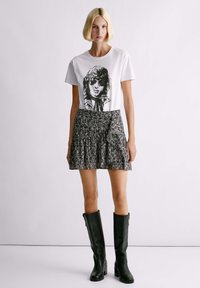 Weißes T-Shirt mit schwarzem Grafikdesign und Text, kombiniert mit einem fließenden, schwarz-weißen gemusterten Minirock und knielangen schwarzen Lederstiefeln.