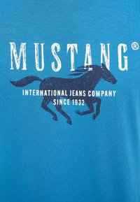 Niebieska koszulka z napisem "MUSTANG" w kolorze białym, z czarną sylwetką konia. Poniżej znajduje się napis "INTERNATIONAL JEANS COMPANY SINCE 1932."