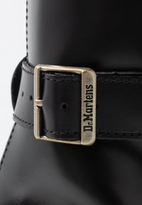 Svarta läderstövlar med en spänne i silverfärg som har det ingraverade namnet "Dr. Martens." Slät yta med förstärkt sömdetalj.