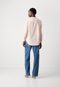 Camicia a maniche lunghe rosa chiaro con orlo curvo, caratterizzata da dettagli a bottoni sul retro, abbinata a jeans in denim blu larghi.